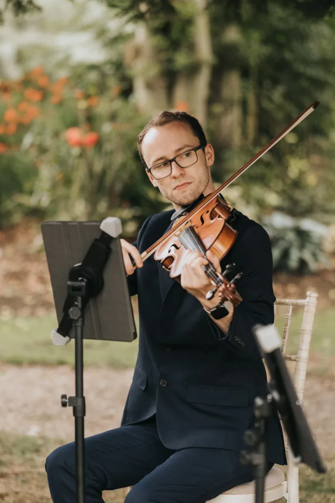 A man in a navy suit and glasses sits outdoors at Hackness Grange, playing a violin. He looks focused, reading sheet music from a tablet on a stand. Green foliage and red flowers create a scenic garden—perfect for any Scarborough wedding photographer. © Aimee Lince Photography - Wedding photographer in Lincolnshire, Yorkshire & Nottinghamshire