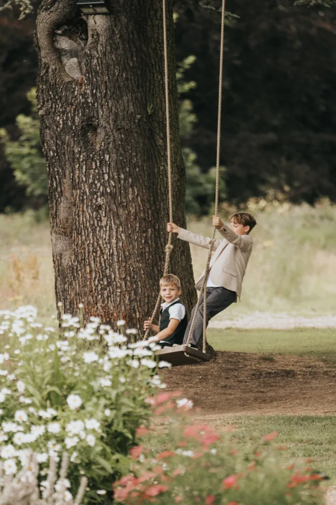 Two young boys play on a rope swing hanging from a large tree at Hackness Grange near Scarborough. One boy sits smiling while the other pushes, surrounded by a grassy field dotted with white daisies and red flowers—a scene perfect for a wedding photographer. © Aimee Lince Photography - Wedding photographer in Lincolnshire, Yorkshire & Nottinghamshire