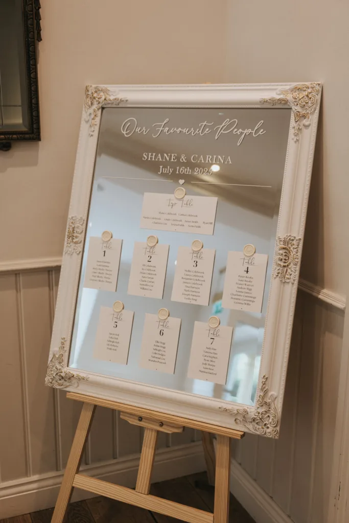 A large ornate white-framed mirror on an easel at Hackness Grange displays a wedding seating chart with "Our Favourite People," names "Shane & Carina," the date "July 16th 2022," and seven table cards, perfect for any Scarborough wedding photographer to capture. © Aimee Lince Photography - Wedding photographer in Lincolnshire, Yorkshire & Nottinghamshire