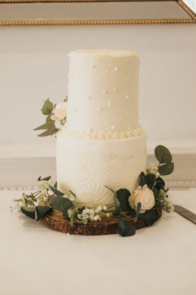 A two-tier white wedding cake sits on a wooden slab at Hackness Grange, Scarborough. The top tier features small white pearls, the bottom has a lace-like pattern, and greenery surrounds the base—perfect for any wedding photographer to capture. © Aimee Lince Photography - Wedding photographer in Lincolnshire, Yorkshire & Nottinghamshire