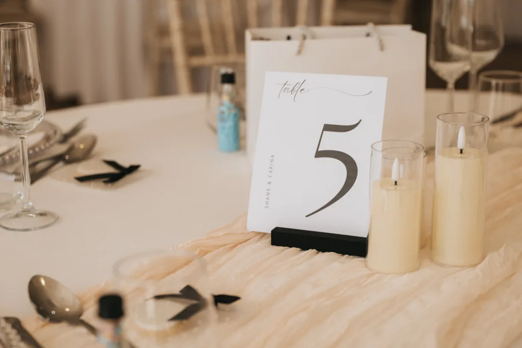 A round table at Hackness Grange in Scarborough is decorated for an event with a “Table 5” card, cream-colored candles, champagne flutes, cutlery, favor bottles, and elegant neutral decor—perfect details for a wedding photographer to capture. © Aimee Lince Photography - Wedding photographer in Lincolnshire, Yorkshire & Nottinghamshire