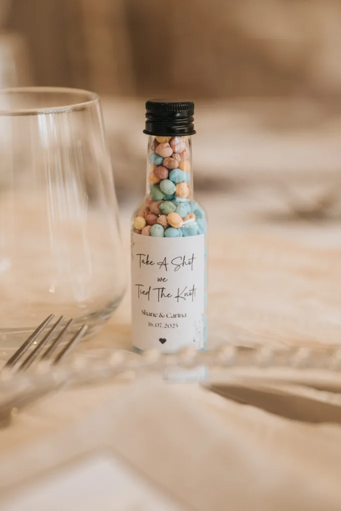 A small glass bottle filled with pastel-colored candy beads sits on a table set at Hackness Grange, Scarborough. The label reads, “Take A Shot we Tied The Knot, Siana & Carina, 16.07.2025,”—perfect for any wedding photographer to capture. © Aimee Lince Photography - Wedding photographer in Lincolnshire, Yorkshire & Nottinghamshire