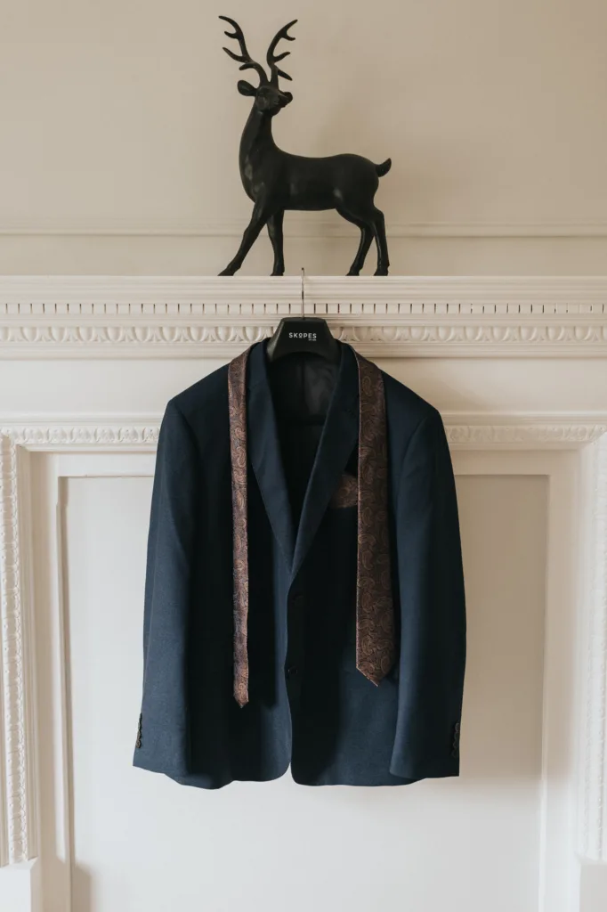 A dark blue suit jacket on a hanger labeled "SERPE" hangs on a white ornate mantel. A patterned brown tie is draped around the collar, ready for a Scarborough wedding photographer to capture. On the mantel sits a decorative black stag statue. © Aimee Lince Photography - Wedding photographer in Lincolnshire, Yorkshire & Nottinghamshire