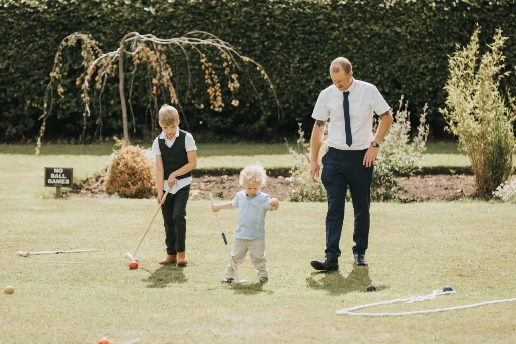 Three people, an adult man and two young boys, play croquet on a grassy lawn at Hackness Grange near Scarborough. The older boy holds a mallet as the toddler steps forward excitedly; nearby, a sign reads "No Ball Games." Perfect for a wedding photographer. © Aimee Lince Photography - Wedding photographer in Lincolnshire, Yorkshire & Nottinghamshire