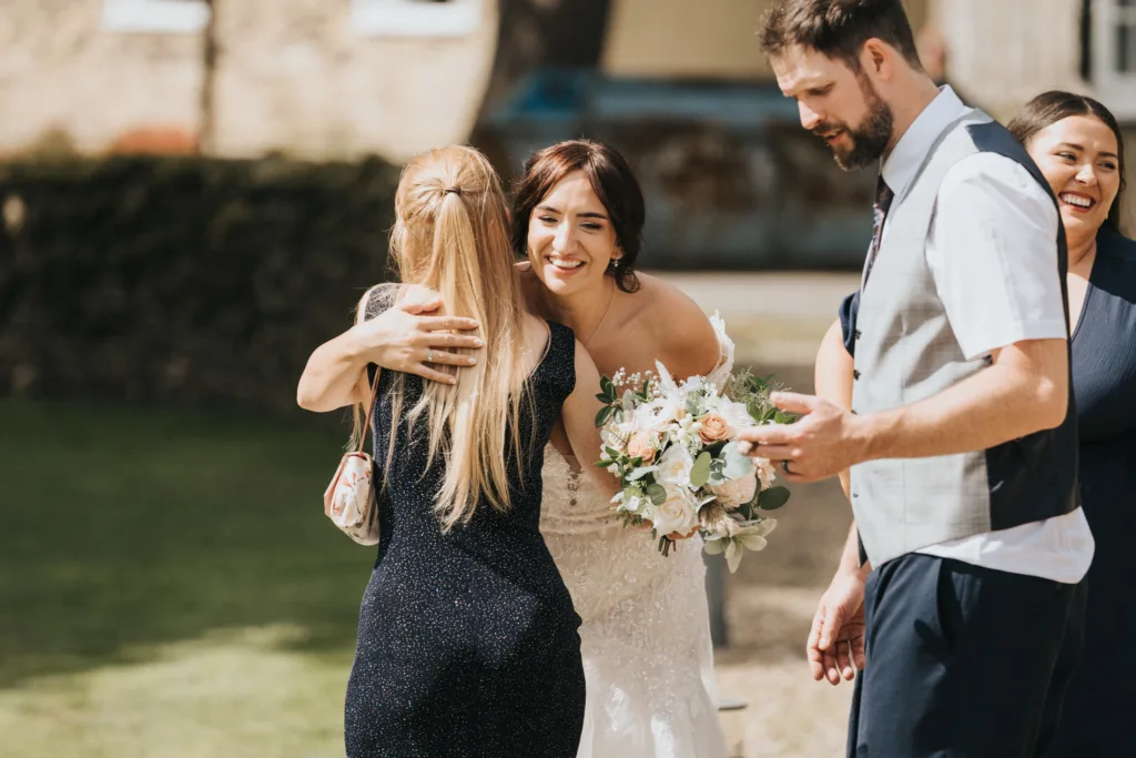 A bride in a white dress hugs a woman in black outdoors at Hackness Grange. The bride, holding a bouquet, smiles warmly as two guests beside her also smile. This sunlit, cheerful moment is perfectly captured by a Scarborough wedding photographer. © Aimee Lince Photography - Wedding photographer in Lincolnshire, Yorkshire & Nottinghamshire