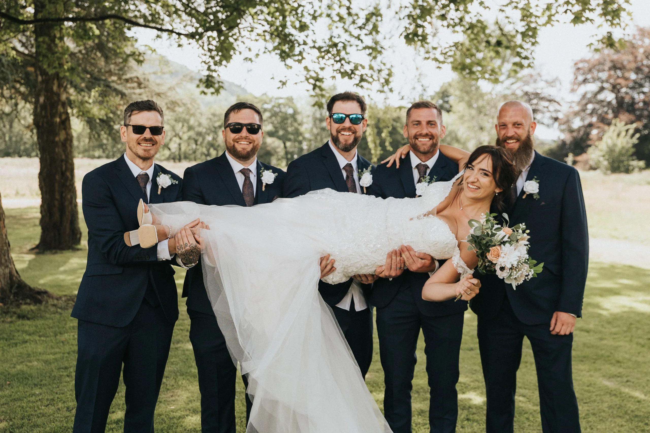 shane-carina-wedding-photography-scarborough-hackness-grange-photographer-copyright-aimee-lince-343-of-649-©-aimee-lince-photography-16-07-2025 Five men in dark suits and sunglasses smile as they hold a bride in a white dress horizontally. Captured by a wedding photographer at Hackness Grange near Scarborough, they pose joyfully on sunlit grass with trees in the background. © Aimee Lince Photography - Wedding photographer in Lincolnshire, Yorkshire & Nottinghamshire