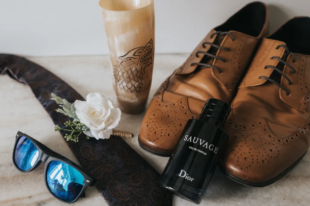 A pair of brown dress shoes, a paisley tie with a white rose boutonnière, blue sunglasses, a Viking-style horn cup with a dragon design, and black Dior Sauvage cologne are arranged on marble—perfect details for any Scarborough wedding photographer. © Aimee Lince Photography - Wedding photographer in Lincolnshire, Yorkshire & Nottinghamshire
