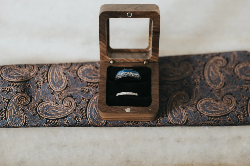 A wooden ring box with a glass lid holds two rings, one dark and one silver, nestled in black velvet. Perfect for a wedding photographer in Scarborough, it rests on blue and bronze paisley fabric atop light marble. © Aimee Lince Photography - Wedding photographer in Lincolnshire, Yorkshire & Nottinghamshire