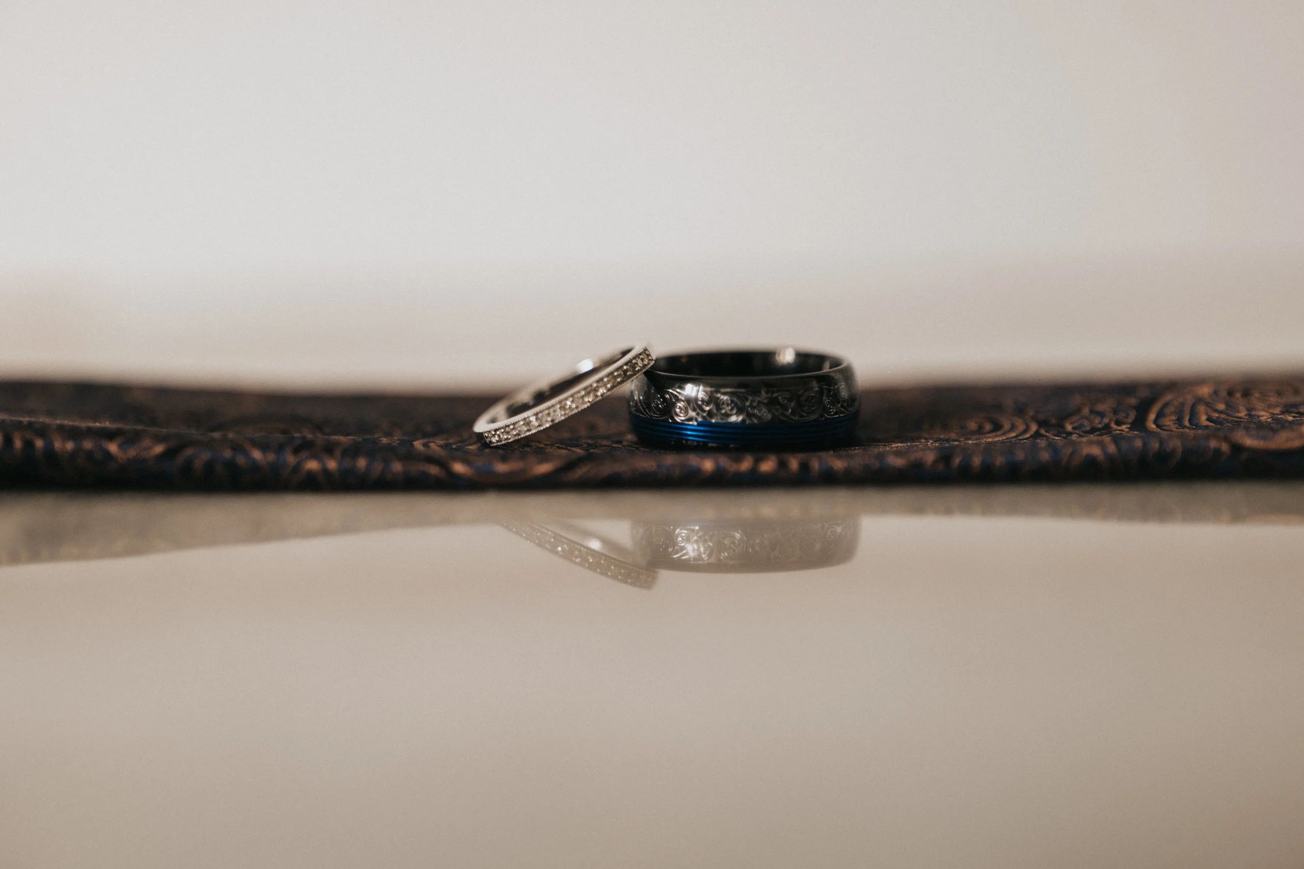 shane-carina-wedding-photography-scarborough-hackness-grange-photographer-copyright-aimee-lince-39-of-649-©-aimee-lince-photography-16-07-2025 Two rings rest on a brown, intricately patterned surface. Captured by a wedding photographer near Hackness Grange, Scarborough, the silver diamond band and darker engraved ring are reflected elegantly against a soft, neutral background. © Aimee Lince Photography - Wedding photographer in Lincolnshire, Yorkshire & Nottinghamshire
