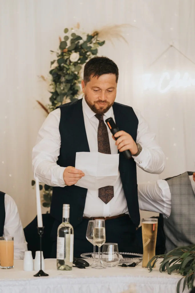 A bearded man in a dark vest and tie stands at a decorated table, holding a microphone and reading from a sheet of paper. Drinks, glassware, and floral decorations hint at a formal event—perhaps captured by a Scarborough wedding photographer at Hackness Grange. © Aimee Lince Photography - Wedding photographer in Lincolnshire, Yorkshire & Nottinghamshire