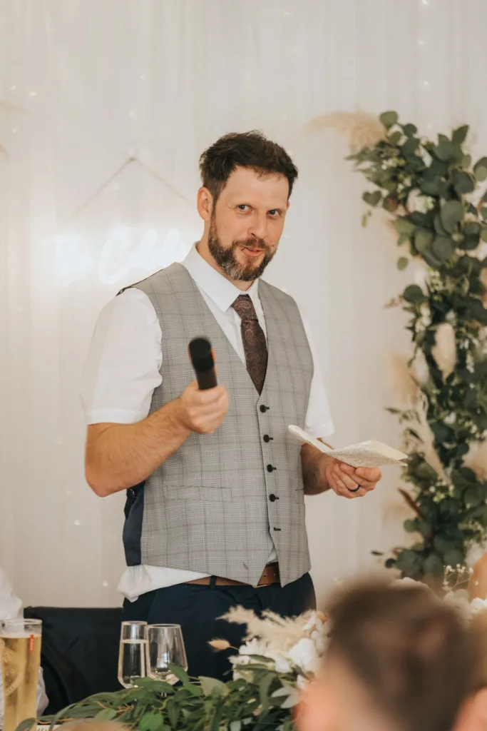 A man with short dark hair and a beard, wearing a gray vest, white shirt, and paisley tie, stands indoors at Hackness Grange holding a microphone and small notepad. He smiles slightly, with greenery and white flowers behind—a scene a wedding photographer would love. © Aimee Lince Photography - Wedding photographer in Lincolnshire, Yorkshire & Nottinghamshire