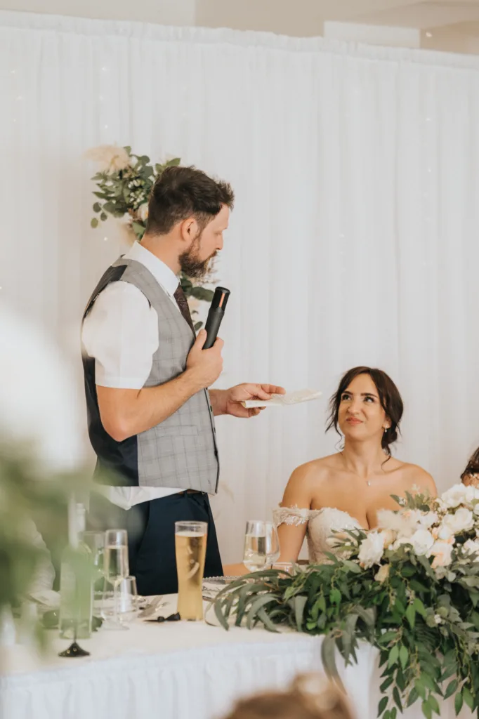 At Hackness Grange near Scarborough, a man in a gray vest gives a speech with a microphone while a woman in an off-shoulder wedding dress looks up at him. The table is adorned with flowers and drinks—an ideal scene for any wedding photographer. © Aimee Lince Photography - Wedding photographer in Lincolnshire, Yorkshire & Nottinghamshire