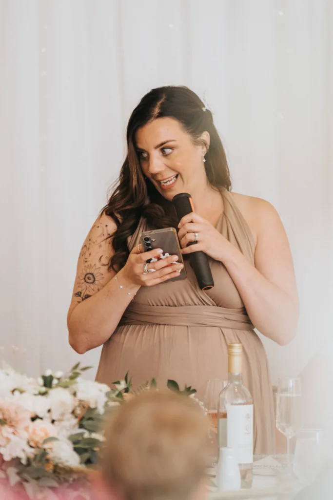 A woman with long brown hair, wearing a taupe dress, smiles while holding a microphone and smartphone. She stands near a table with flowers, wine, and glasses. Her arm has a tattoo, framed by a white curtain—perfect for a Hackness Grange wedding photographer in Scarborough. © Aimee Lince Photography - Wedding photographer in Lincolnshire, Yorkshire & Nottinghamshire