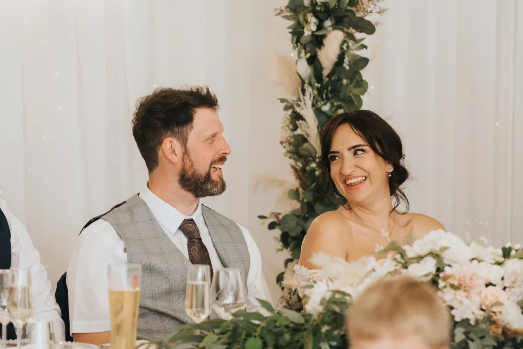 A man in a vest and tie and a woman in a white, off-the-shoulder dress smile at each other at Hackness Grange. Captured by a wedding photographer near Scarborough, they sit at a flower-adorned table with champagne glasses and a floral arch behind them. © Aimee Lince Photography - Wedding photographer in Lincolnshire, Yorkshire & Nottinghamshire