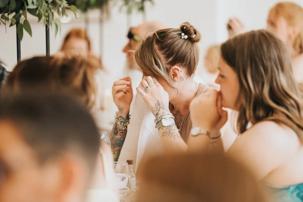 A woman with tattoos and jewelry wipes her eyes with a napkin at a table, surrounded by guests. The emotional scene, framed by greenery, captures a heartfelt moment—perfect for a wedding photographer at Hackness Grange near Scarborough. © Aimee Lince Photography - Wedding photographer in Lincolnshire, Yorkshire & Nottinghamshire