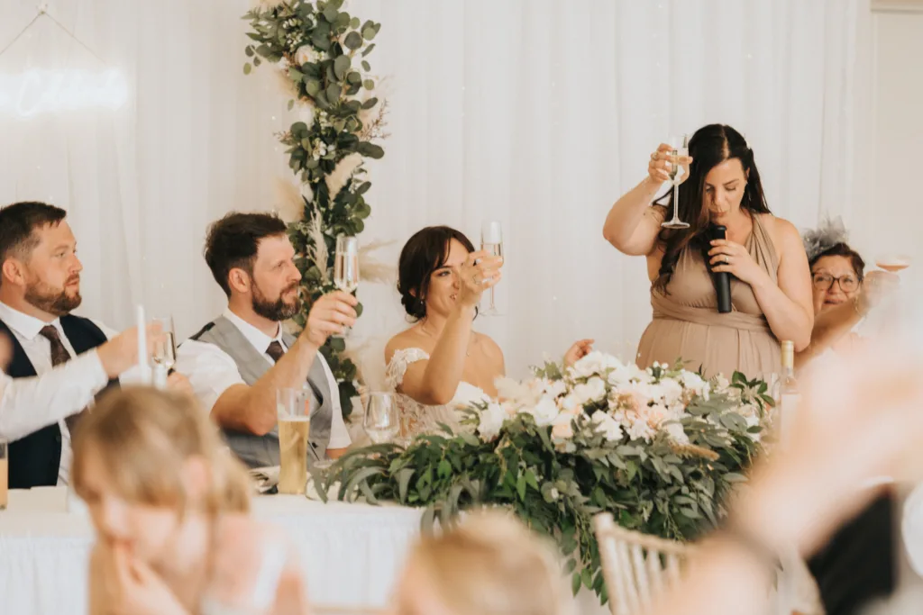 A wedding party sits at a decorated table with greenery and white flowers at Hackness Grange. The bride and groom, among others, raise champagne glasses in a toast as a woman in beige speaks into a microphone. A Scarborough wedding photographer captures the celebratory mood. © Aimee Lince Photography - Wedding photographer in Lincolnshire, Yorkshire & Nottinghamshire