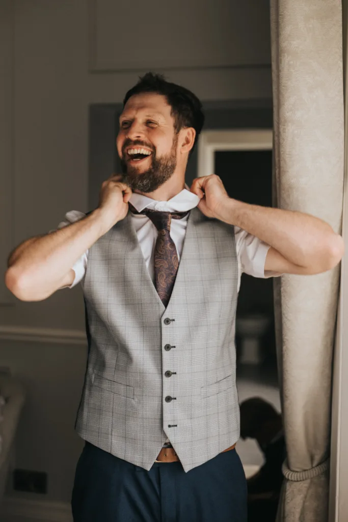 A smiling man with a beard adjusts his brown patterned tie. He wears a white shirt and gray plaid vest, standing near a light curtain in a softly lit room at Hackness Grange, Scarborough—appearing happy and relaxed, perfect for a wedding photographer’s lens. © Aimee Lince Photography - Wedding photographer in Lincolnshire, Yorkshire & Nottinghamshire
