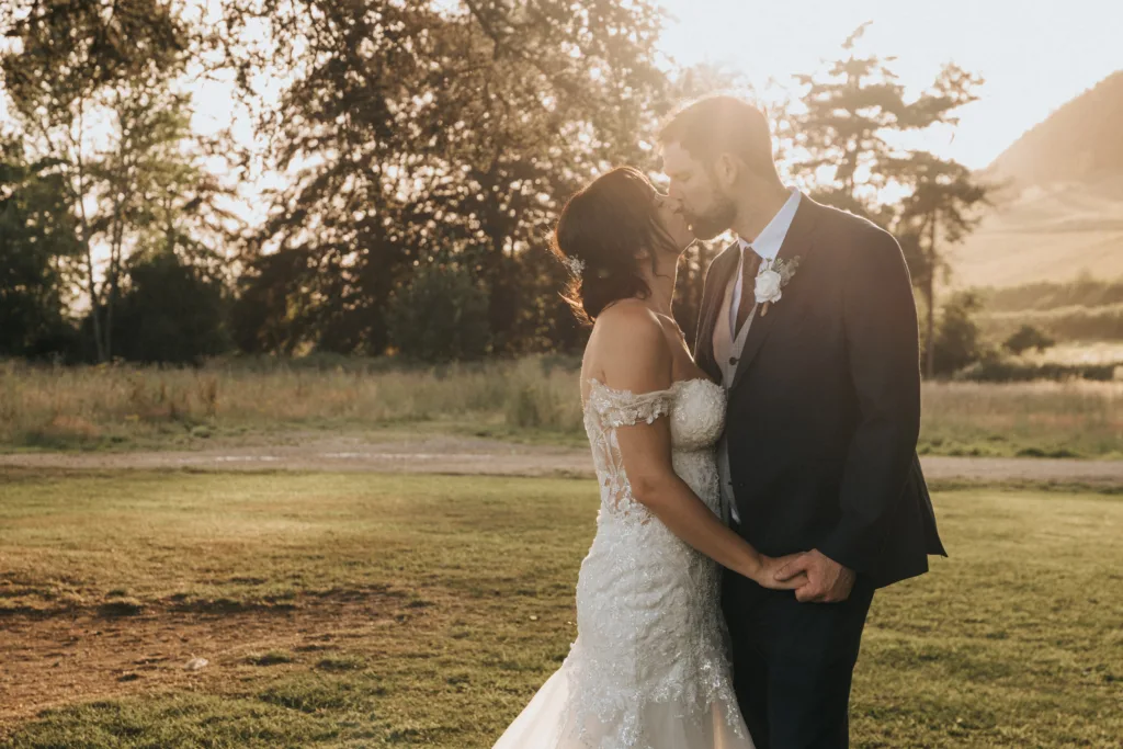 A bride in a white lace dress and a groom in a dark suit kiss outdoors at sunset at Hackness Grange, holding hands on a grassy field, with sunlight streaming through trees—a perfect moment captured by a Scarborough wedding photographer. © Aimee Lince Photography - Wedding photographer in Lincolnshire, Yorkshire & Nottinghamshire