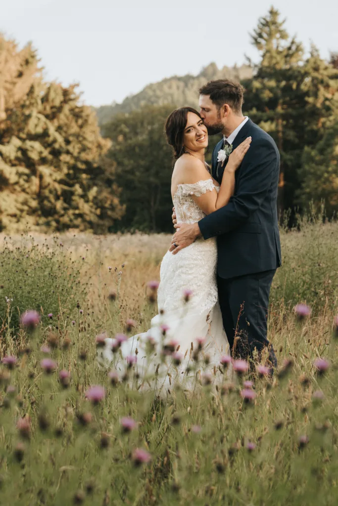 A bride in an off-the-shoulder white gown and a groom in a dark suit embrace and smile in a field of purple wildflowers, captured by a Scarborough wedding photographer near Hackness Grange, with hills and trees under a clear sky. © Aimee Lince Photography - Wedding photographer in Lincolnshire, Yorkshire & Nottinghamshire