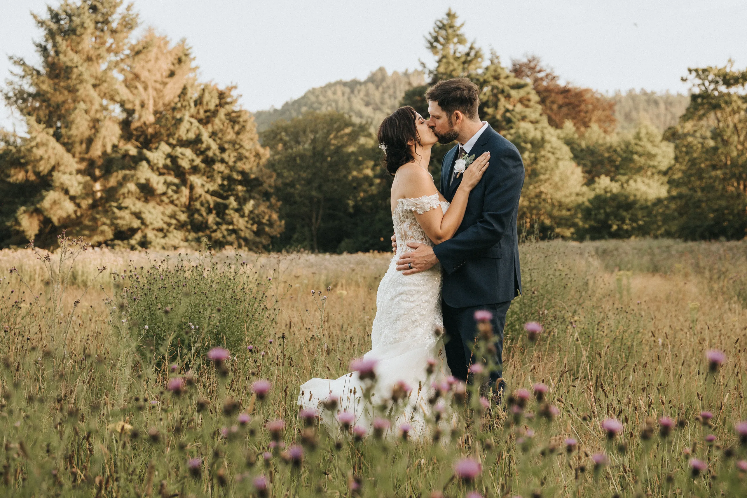 shane-carina-wedding-photography-scarborough-hackness-grange-photographer-copyright-aimee-lince-520-of-649-©-aimee-lince-photography-16-07-2025 A bride in a white, off-shoulder gown and a groom in a dark suit kiss in a field of wildflowers at Hackness Grange near Scarborough, beautifully captured by their wedding photographer under a clear sky amid romantic greenery. © Aimee Lince Photography - Wedding photographer in Lincolnshire, Yorkshire & Nottinghamshire