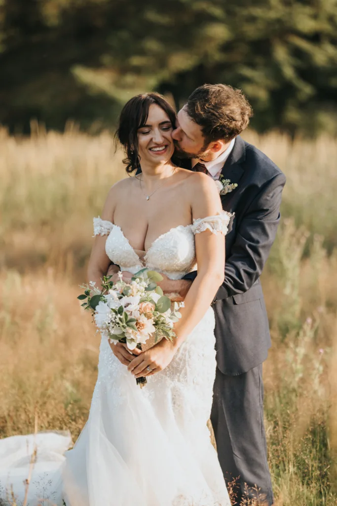 A bride in a white off-shoulder gown holds a bouquet in a grassy field at Hackness Grange, smiling as the groom embraces her from behind. Captured by a wedding photographer, the warm scene glows with romantic, soft sunlight. © Aimee Lince Photography - Wedding photographer in Lincolnshire, Yorkshire & Nottinghamshire