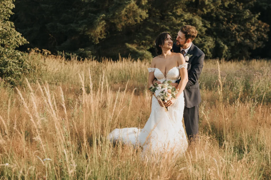 A joyful bride in a white off-shoulder gown holds flowers and smiles as her groom hugs her from behind in a sunlit field at Hackness Grange. Captured by a Scarborough wedding photographer, the couple looks relaxed amid green trees and tall grass. © Aimee Lince Photography - Wedding photographer in Lincolnshire, Yorkshire & Nottinghamshire