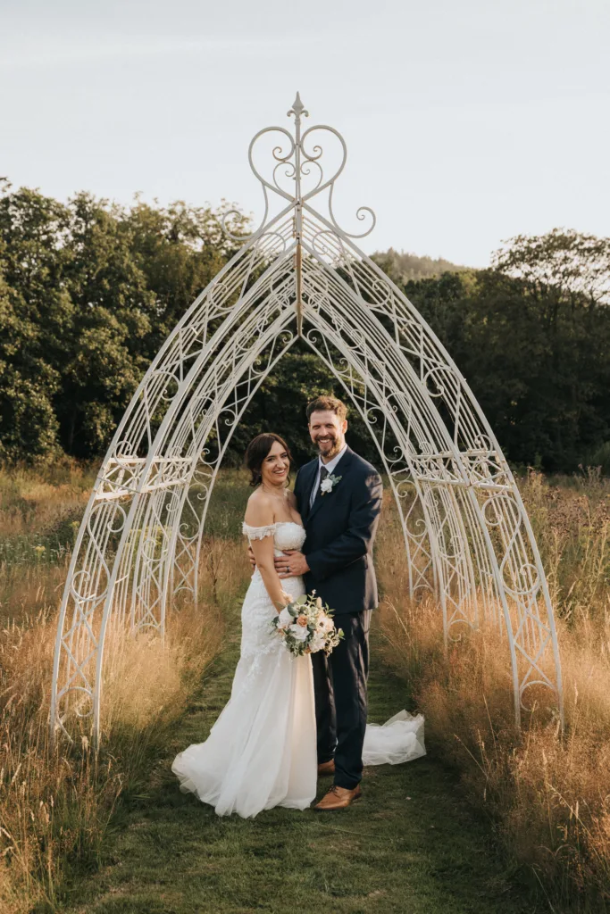 A bride in an off-shoulder white gown and a groom in a navy suit stand smiling under a decorative archway at Hackness Grange, surrounded by tall grass and trees. Captured by a Scarborough wedding photographer, the bride holds a bouquet of white flowers. © Aimee Lince Photography - Wedding photographer in Lincolnshire, Yorkshire & Nottinghamshire