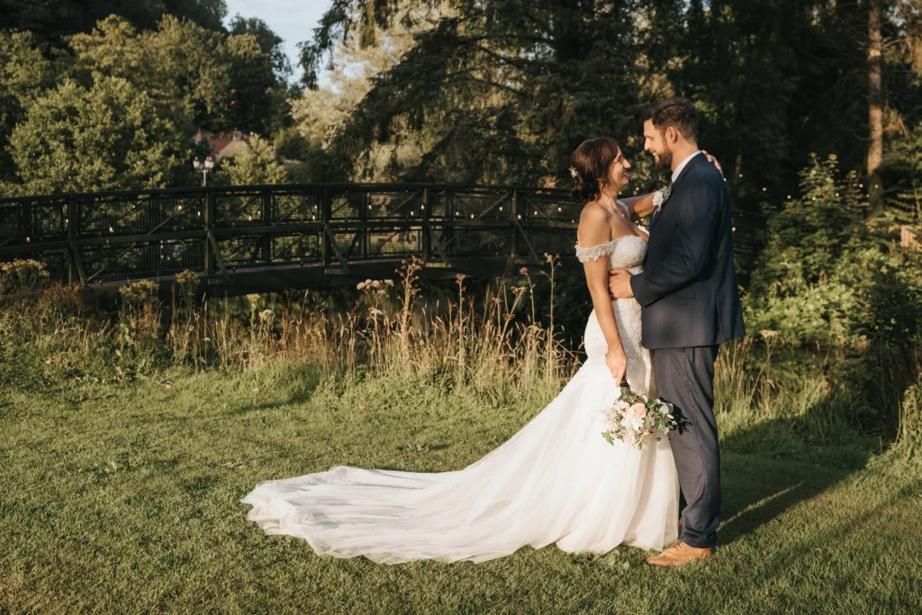 A bride in a white gown holding a bouquet and a groom in a navy suit stand smiling at each other in the sunlit gardens of Hackness Grange, Scarborough, with lush greenery and a black bridge behind them—perfect for any wedding photographer. © Aimee Lince Photography - Wedding photographer in Lincolnshire, Yorkshire & Nottinghamshire