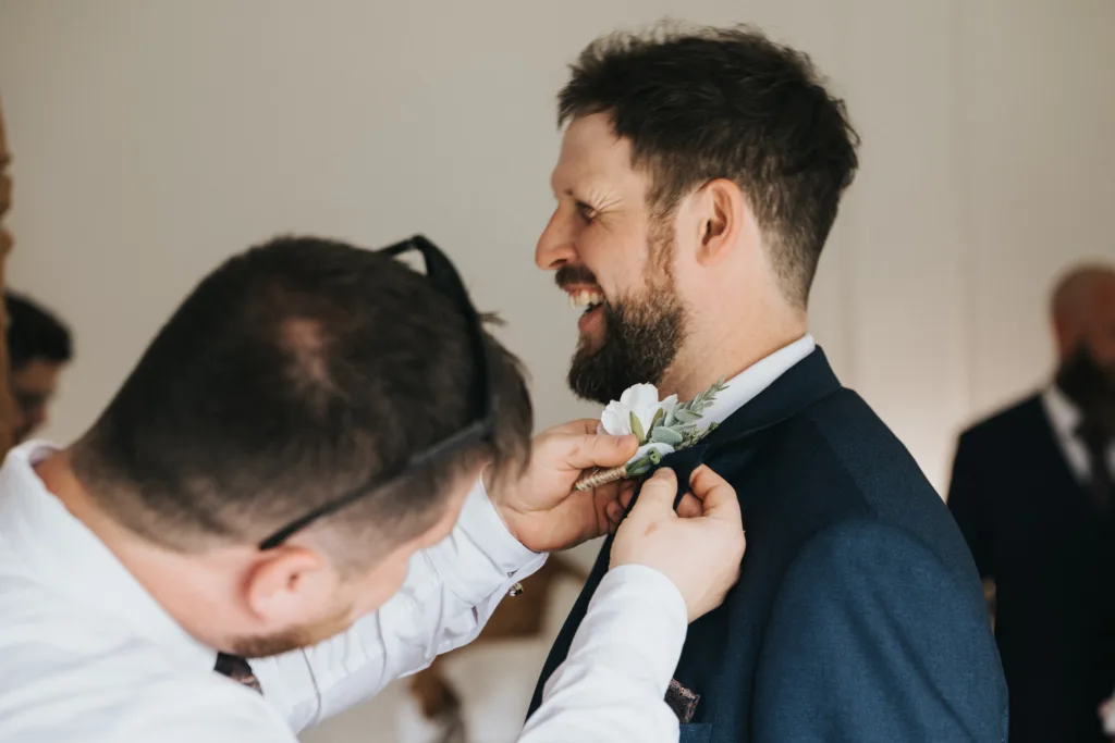 A smiling man in a suit has a white boutonniere pinned to his lapel by another man wearing glasses. Both are indoors, preparing for a special event—likely a wedding at Hackness Grange, beautifully captured by a talented wedding photographer. © Aimee Lince Photography - Wedding photographer in Lincolnshire, Yorkshire & Nottinghamshire