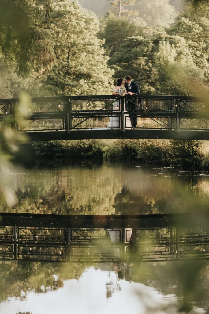 A couple in wedding attire stands closely together on a metal bridge over a calm lake at Hackness Grange, near Scarborough. Their embrace and the bridge are mirrored in the water below, creating a serene scene for any wedding photographer to capture. © Aimee Lince Photography - Wedding photographer in Lincolnshire, Yorkshire & Nottinghamshire