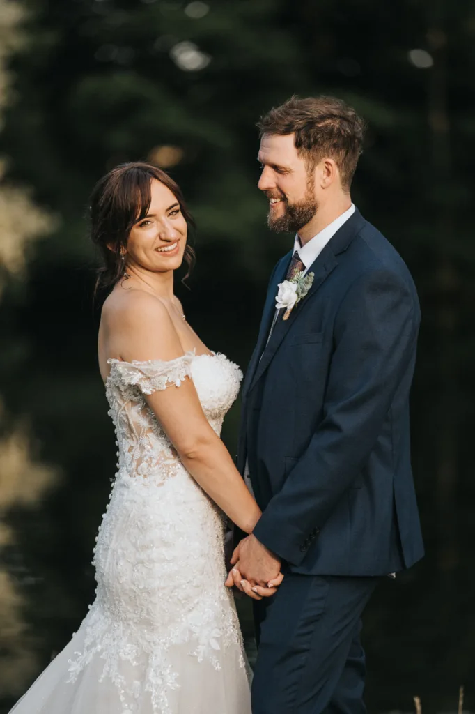 A bride in an off-the-shoulder lace wedding dress and a groom in a navy suit with a boutonnière stand outdoors at Hackness Grange, holding hands and smiling at each other—captured by a Scarborough wedding photographer against a blurred green backdrop. © Aimee Lince Photography - Wedding photographer in Lincolnshire, Yorkshire & Nottinghamshire