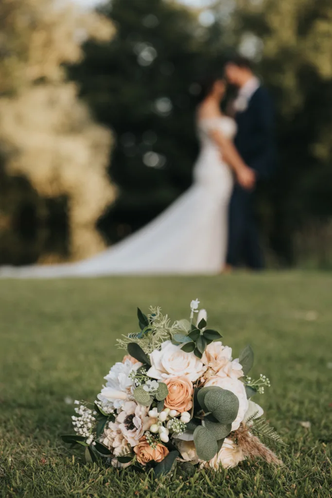A bridal bouquet with white and peach flowers and greenery lies on grass in sharp focus at Hackness Grange; in the blurred background, a wedding photographer captures the bride and groom standing close together outdoors amid trees. © Aimee Lince Photography - Wedding photographer in Lincolnshire, Yorkshire & Nottinghamshire