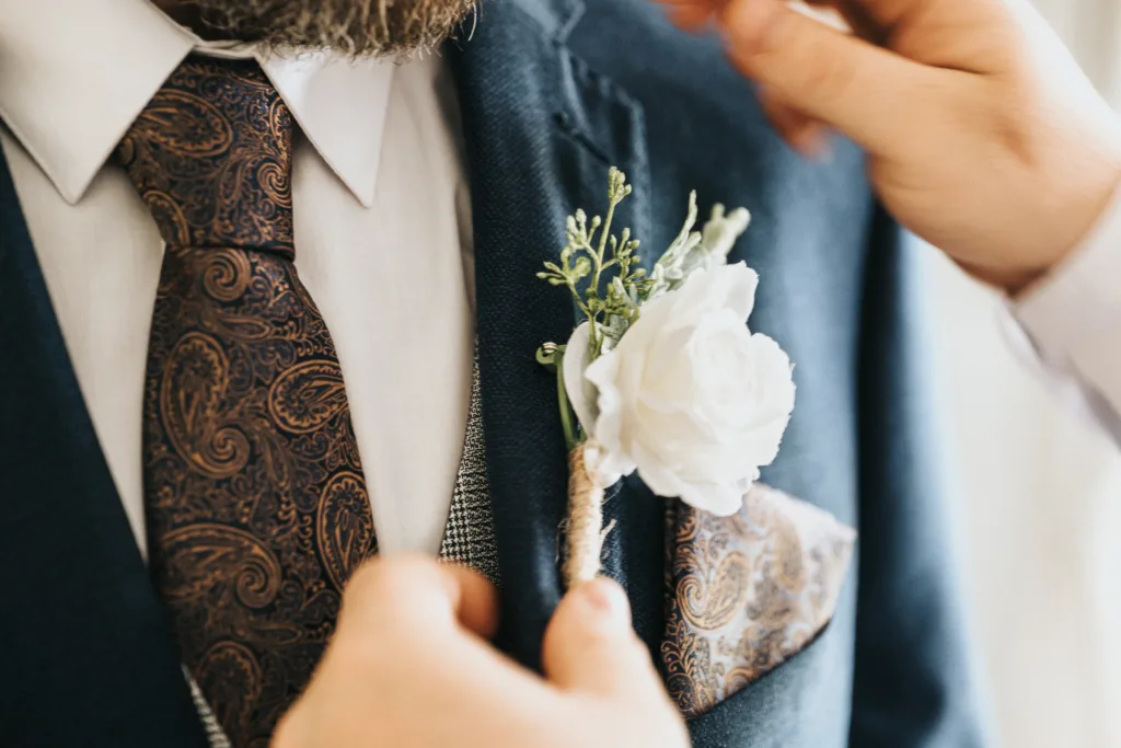 Close-up of a man in a navy blue suit with a brown paisley tie and pocket square as someone pins a white rose boutonniere to his lapel. Captured by a Hackness Grange wedding photographer, the focus is on the flower and suit details. © Aimee Lince Photography - Wedding photographer in Lincolnshire, Yorkshire & Nottinghamshire