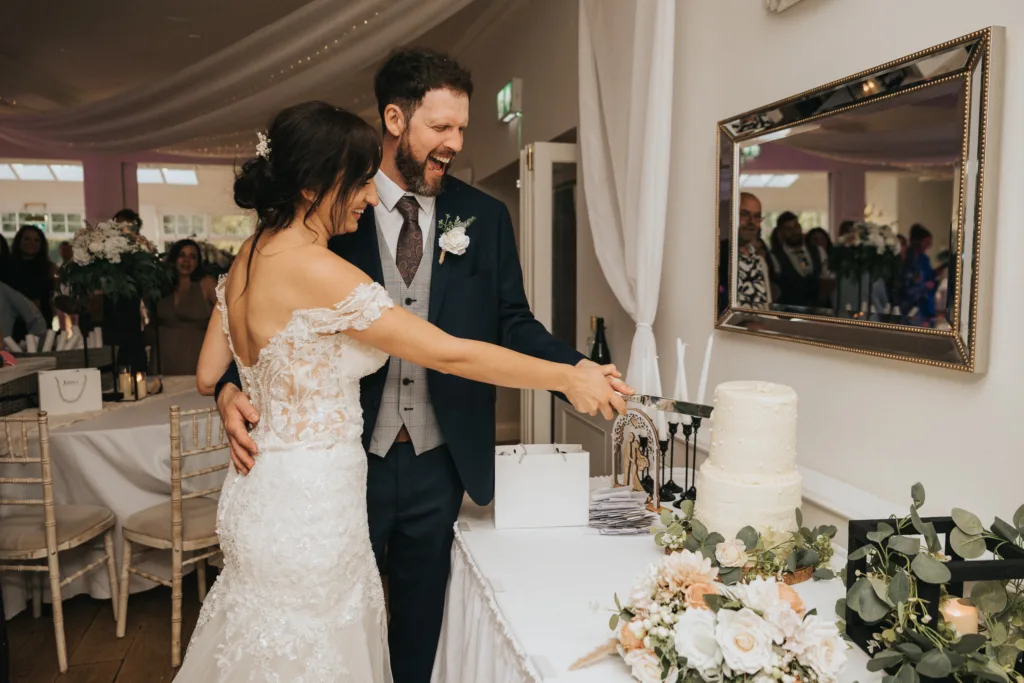 A bride and groom smile joyfully as they cut a tiered white wedding cake at their Hackness Grange celebration in Scarborough. Flowers and candles adorn the table, while a wedding photographer captures every cherished moment. © Aimee Lince Photography - Wedding photographer in Lincolnshire, Yorkshire & Nottinghamshire