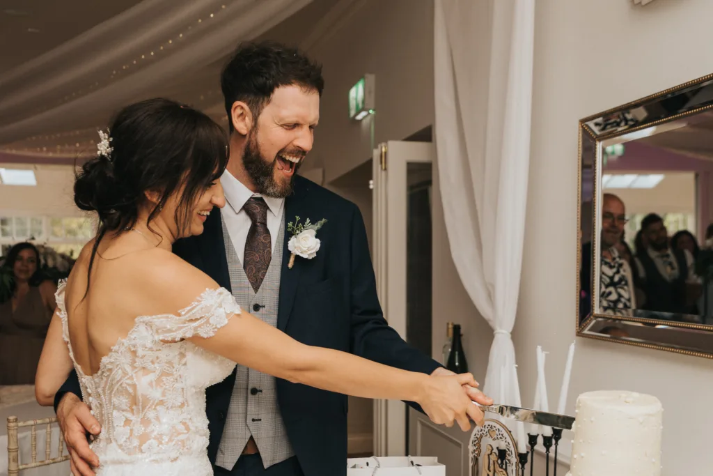 A bride and groom, both smiling joyfully, cut a white wedding cake together at Hackness Grange. The bride wears an off-shoulder lace gown, and the groom is in a dark suit. Guests and a large mirror appear in the softly lit background. © Aimee Lince Photography - Wedding photographer in Lincolnshire, Yorkshire & Nottinghamshire