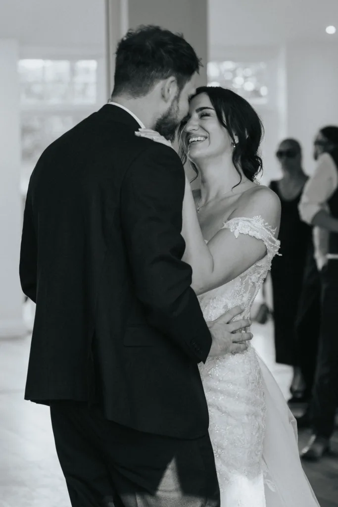 A bride and groom share a joyful moment while dancing at Hackness Grange. The groom wears a dark suit and the bride an off-the-shoulder white gown. Captured by a wedding photographer, they smile lovingly in this softly lit, black and white photo. © Aimee Lince Photography - Wedding photographer in Lincolnshire, Yorkshire & Nottinghamshire