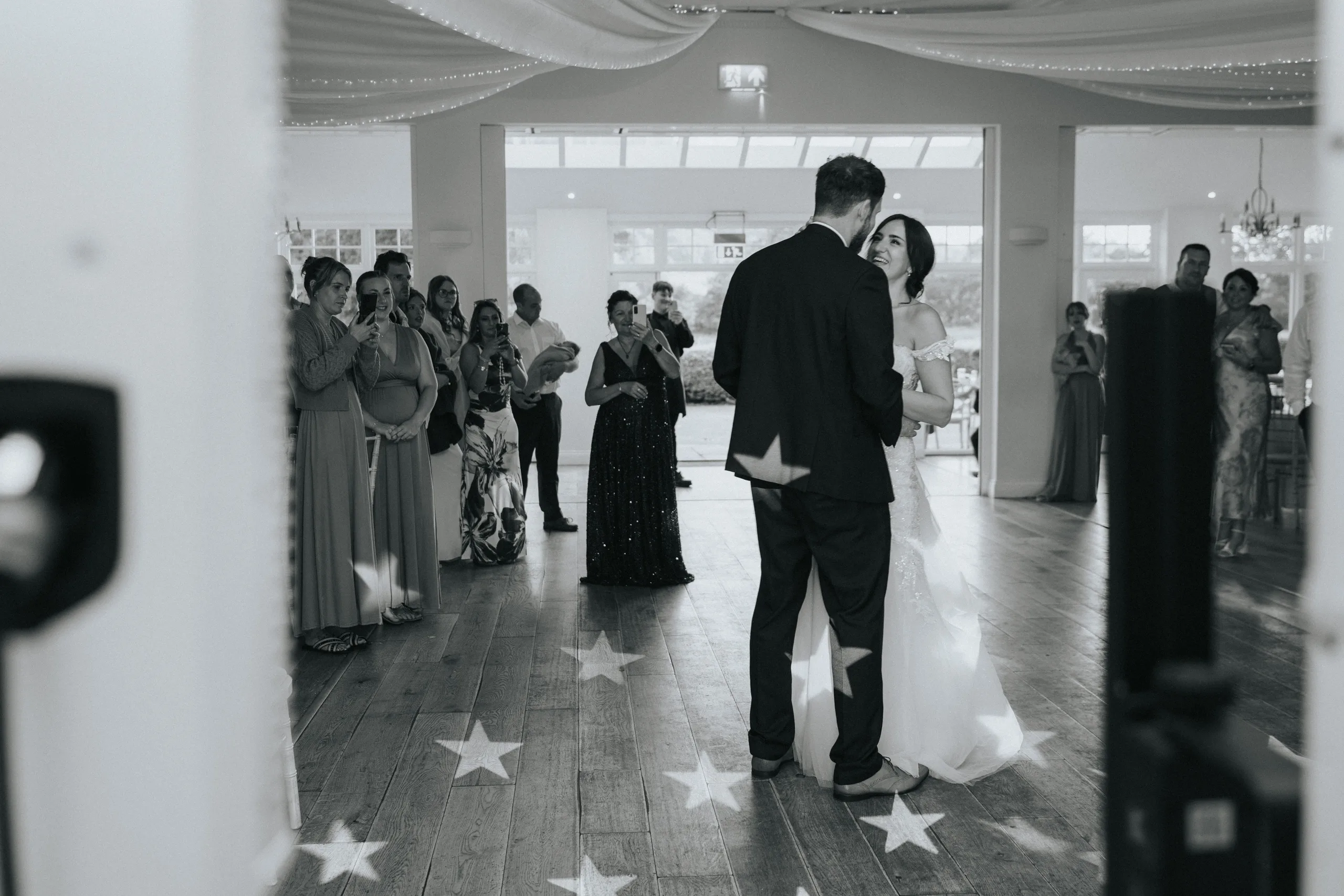 shane-carina-wedding-photography-scarborough-hackness-grange-photographer-copyright-aimee-lince-578-of-649-©-aimee-lince-photography-16-07-2025 A bride and groom share their first dance in a bright, elegant venue at Hackness Grange near Scarborough as guests watch and smile. The wooden floor is decorated with star-shaped lights, captured by a wedding photographer in timeless black and white. © Aimee Lince Photography - Wedding photographer in Lincolnshire, Yorkshire & Nottinghamshire