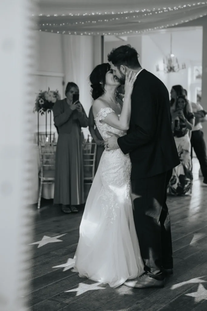 A bride and groom share a close, joyful dance in a softly lit room at Hackness Grange, surrounded by guests. The bride wears a lace wedding gown and the groom a dark suit, as decorative lights and star patterns create magic for their Scarborough wedding photographer. © Aimee Lince Photography - Wedding photographer in Lincolnshire, Yorkshire & Nottinghamshire