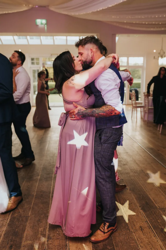 A couple embraces while dancing on a wooden floor scattered with star-shaped light projections at Hackness Grange near Scarborough. Captured by a wedding photographer, guests in formal attire dance in the softly lit room. © Aimee Lince Photography - Wedding photographer in Lincolnshire, Yorkshire & Nottinghamshire