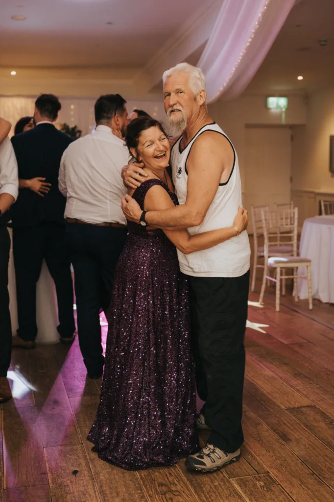 An older couple dances and smiles warmly at a festive indoor event near Scarborough. On the wooden floor of Hackness Grange, they embrace—she in a sparkly purple dress, he in a tank top and hiking shoes—amid joyful guests and decorated tables, ideal for any wedding photographer. © Aimee Lince Photography - Wedding photographer in Lincolnshire, Yorkshire & Nottinghamshire