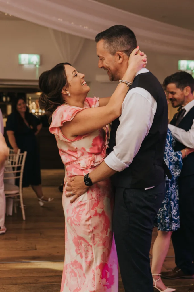 A woman in a pink floral dress and a man in a white shirt and vest smile and embrace while dancing at an indoor event. Captured by a Hackness Grange wedding photographer near Scarborough, the joyful atmosphere shines among guests and white chairs. © Aimee Lince Photography - Wedding photographer in Lincolnshire, Yorkshire & Nottinghamshire