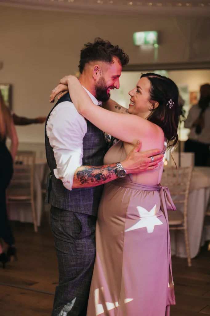 A bearded man in a grey plaid suit and white shirt smiles while dancing closely with a woman in a mauve dress at Hackness Grange, Scarborough. Captured by a wedding photographer, they embrace warmly, making eye contact in the softly lit room. © Aimee Lince Photography - Wedding photographer in Lincolnshire, Yorkshire & Nottinghamshire