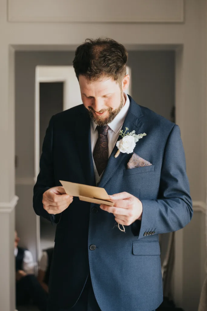 A man in a navy suit with a floral boutonniere and patterned tie smiles while reading a card indoors at Hackness Grange. He stands in a softly lit room with neutral tones—a wedding photographer in Scarborough captures the moment. © Aimee Lince Photography - Wedding photographer in Lincolnshire, Yorkshire & Nottinghamshire