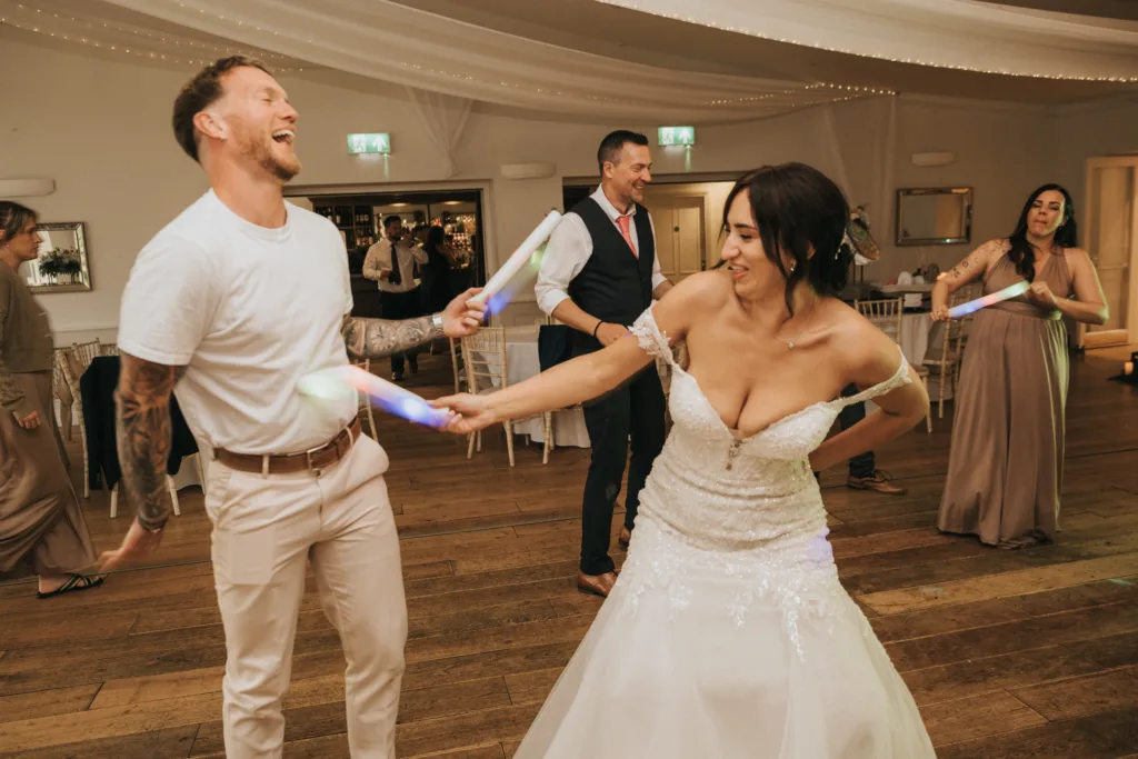 A bride in a white wedding dress playfully dances with a man in casual clothes at a reception. Both are laughing and holding glowing sticks, while guests in formal attire smile and dance—captured by a skilled wedding photographer in Scarborough’s Hackness Grange. © Aimee Lince Photography - Wedding photographer in Lincolnshire, Yorkshire & Nottinghamshire