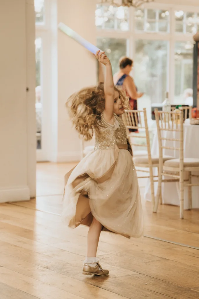 A young girl in a sparkly beige dress twirls joyfully on the wooden floor at Hackness Grange, her light-up wand held high. Captured by a Scarborough wedding photographer, her curls flow as she spins in the sunlit room with white chairs and tables behind her. © Aimee Lince Photography - Wedding photographer in Lincolnshire, Yorkshire & Nottinghamshire