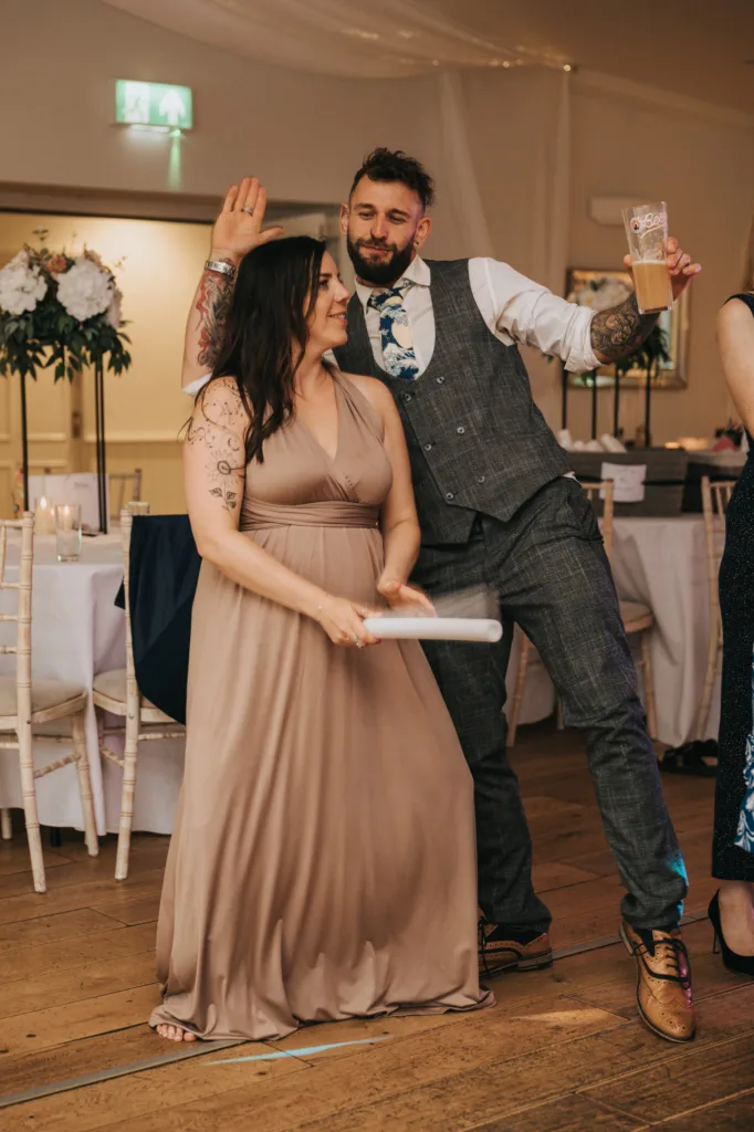 A woman in a taupe dress holds a rolled-up paper, smiling at a man beside her. The man, in a gray checked suit and patterned tie, poses playfully with a drink. Captured by a Scarborough wedding photographer at Hackness Grange’s festive indoor event. © Aimee Lince Photography - Wedding photographer in Lincolnshire, Yorkshire & Nottinghamshire