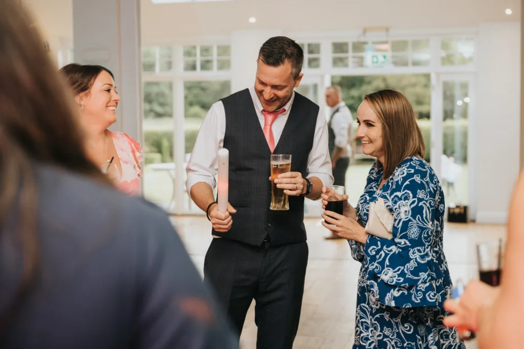 A man in a suit vest, holding a drink and a long object, smiles and interacts with two women at Hackness Grange. Captured by a wedding photographer, they enjoy a friendly conversation at this Scarborough indoor event. © Aimee Lince Photography - Wedding photographer in Lincolnshire, Yorkshire & Nottinghamshire