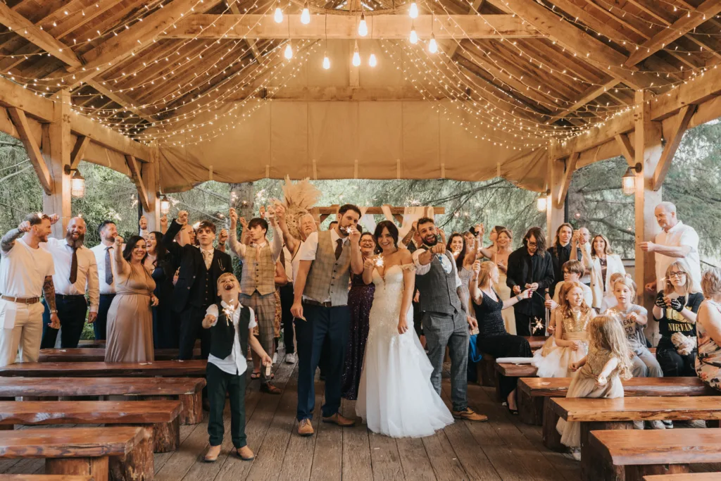 A joyful wedding group poses under a wooden pavilion with string lights at Hackness Grange, Scarborough. The wedding photographer captures the bride, groom, and cheering guests as they smile and laugh, creating a lively, celebratory atmosphere. © Aimee Lince Photography - Wedding photographer in Lincolnshire, Yorkshire & Nottinghamshire