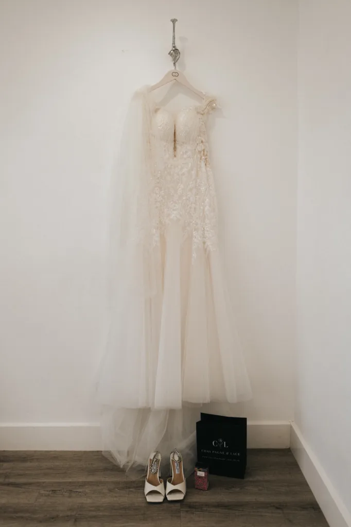 An elegant white lace wedding dress hangs on a white hanger against a plain wall at Hackness Grange. Below, white high-heeled shoes, a black gift bag, and a small red box sit neatly—perfect details for any Scarborough wedding photographer. © Aimee Lince Photography - Wedding photographer in Lincolnshire, Yorkshire & Nottinghamshire