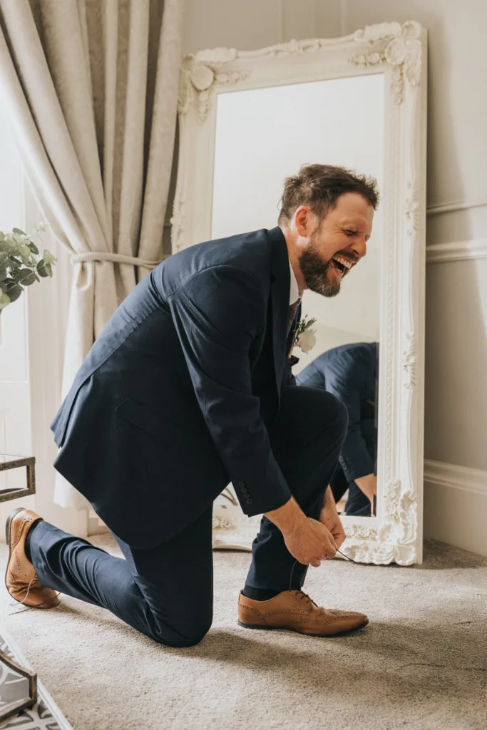 A man in a navy suit and brown dress shoes kneels on one knee, laughing as he ties his shoe. Captured by a Scarborough wedding photographer, he’s indoors at Hackness Grange, in front of an ornate white mirror and elegant decor. © Aimee Lince Photography - Wedding photographer in Lincolnshire, Yorkshire & Nottinghamshire